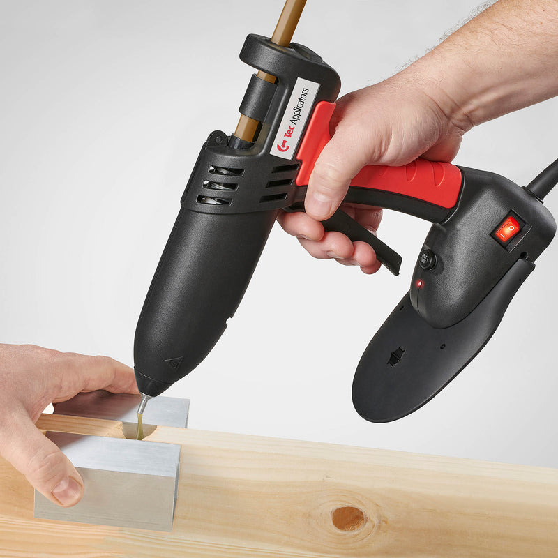 Person using a Power Adhesives 830-12 glue gun to apply hot melt adhesive between two aluminum heat sync blocks on a wooden surface.